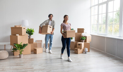 Fototapeta na wymiar Satisfied happy millennial caucasian male and female carry cardboard boxes, enjoy moving in empty apartment