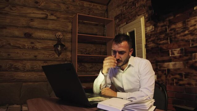 A distressed businessman clutches his head, waves his hands, got losses. A young man with a beard works at home using a laptop. Business concept remotely, online. 4K UHD.