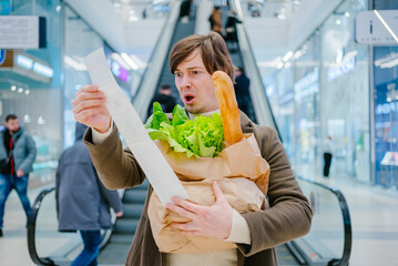 Man in a coat holds a paper check and a bag with a baguette and lettuce in a shopping mall and is...