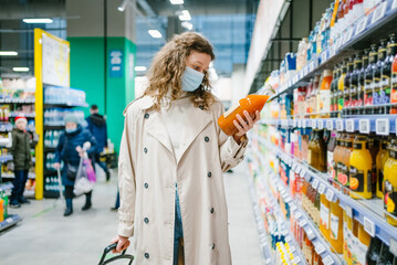 Young curly woman in a medical mask and a beige raincoat chooses juice in a glass bottle in a...