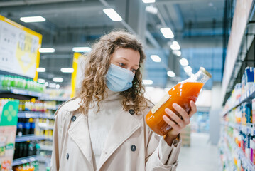 Young curly woman in a medical mask and a beige raincoat chooses juice in a glass bottle in a grocery supermarket, close-up