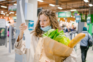 Young woman in a medical mask looks shocked at a paper check in a grocery supermarket holding a...