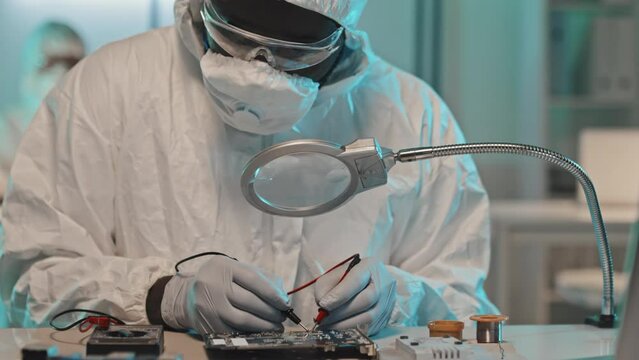 Tilt down shot of electronics engineer in coveralls, mask and gloves using multimeter while testing computer circuit board in laboratory
