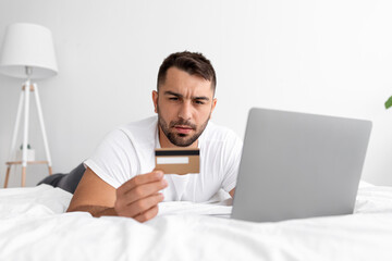 Serious young muscular european male hold credit card near laptop lies on bed and shopping online in bedroom