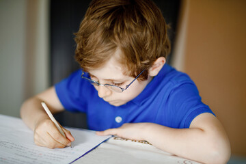 Naklejka na ściany i meble Portrait of little kid boy with glasses at home making homework, writing letters with colorful pens. Little child doing exercise, indoors. Elementary school and education, home schooling concept.