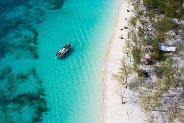 Top view of lonely wooden boat among turquoise water of ocean, thin strip of the white sandy beach and greenery