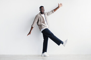 Full length portrait of funny young black guy in casual wear walking against white studio wall,...