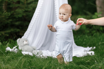 Cute little baby boy takes the first steps on the grass in summer, on a Sunny day, playing in green garden. Selective focus, space for text. happy childhood