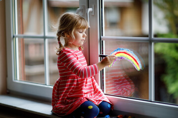Adoralbe little toddler girl with rainbow painted with colorful window color during pandemic...