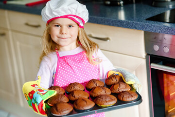 Cute little toddler girl holding fresh baked homemade chocolate muffins cupcakes at home indoors. Adorable preschool blond child with apron with bunny and carrot cookie in domestic kitchen.