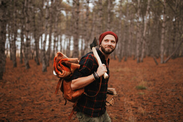 Bearded millennial man in plaid shirt is hiking with backpack, axe and firewood like lumberjack.