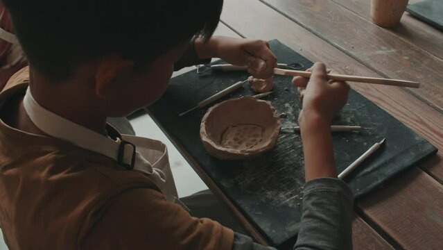 Over-shoulder of little Asian boy sitting at table in pottery school, carving pattern on inside of bowl made of raw clay, learning pottery