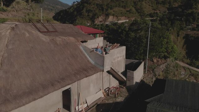 Aerial flying away from three men standing on the deck top floor of unfinished building house in a third world country Kabayan, Benguet, Philippines revealing the entire village