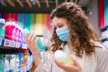 Portrait of woman shopper with curly hair wearing medical mask in supermarket chooses shampoo during coronavirus outbreak