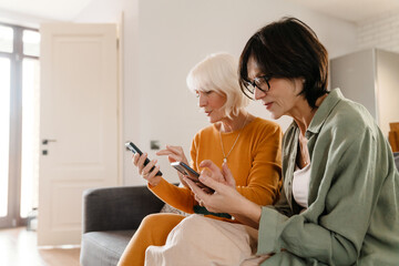 Mature two women using cellphones together while sitting on sofa