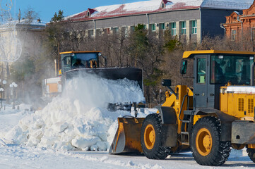Clearing the road from snow. Two excavator tractors are working on a city street after a snowfall. Snow dust from the bucket of the machine flies in the wind. Winter sunny day.