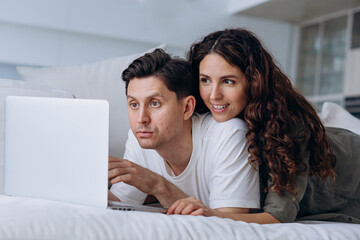 Fototapeta premium Joyful young couple of wife and brunet husband discusses chosen online purchases at sell-out via laptop lying on soft sofa in living room