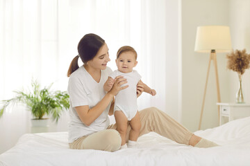 Obraz na płótnie Canvas Portrait of a happy mother with her child on the bed at home. Beautiful young mom sitting on the bed and holding her cute baby boy wearing comfy white cotton onesies. Family, care, and comfort concept