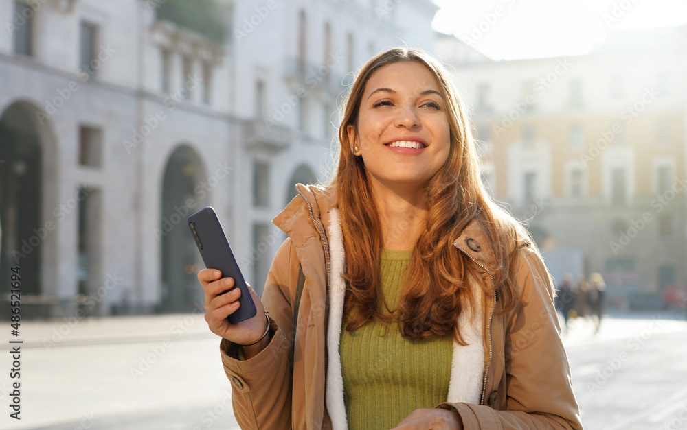 Wall mural Portrait of beautiful young woman holding smartphone on city background looking to the side and staring away thinking