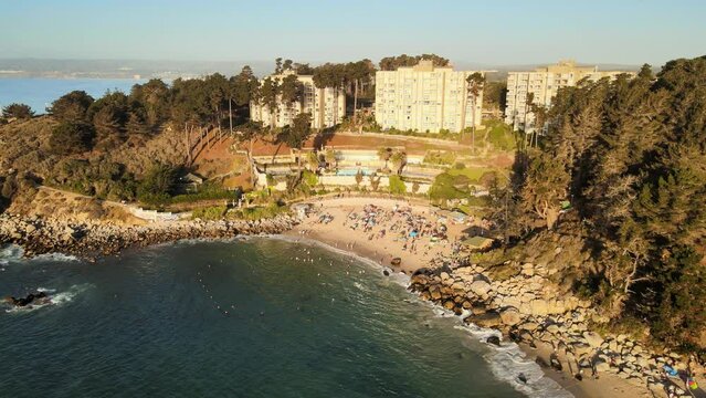 Aerial dolly out of people in sand shore near turquoise sea surrounded by pine woods at sunset, El Canelillo beach, Chile