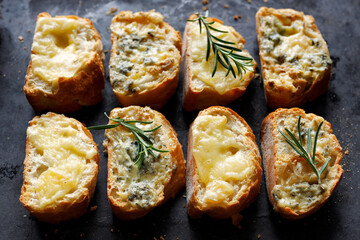 Crostini, cheese toasts  of sliced baguette with two types of cheese and fresh rosemary on a dark background, top view.