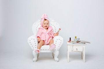 A cute little baby girl in a pink terry dressing gown with a chair and a mini dressing table with cosmetics is isolated on a white background, repeats the behavior of her mother