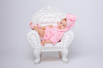 A cute little baby girl in a pink terry dressing gown with a chair and a mini dressing table with...