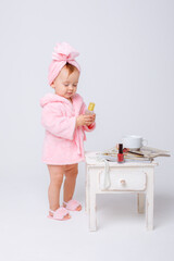 A cute little baby girl in a pink terry dressing gown with a mini dressing table and cosmetics is...