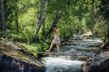 Young woman in a summer dress walking on a wooden bridge over mountain stream. Excursion in nature reserve. Picturesque forest landscape.