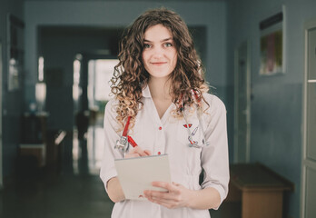A woman doctor in a white coat stands against the background of a polyclinic and holds a pen and a notebook in her hands.