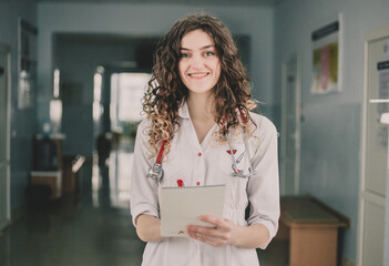 A woman doctor in a white coat stands against the background of a polyclinic and holds a pen and a notebook in her hands.