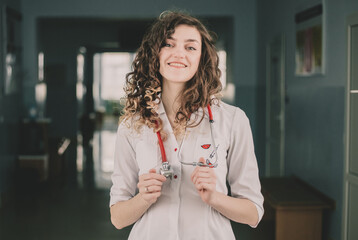 A woman doctor in a protective mask and a white coat stands against the backdrop of a polyclinic