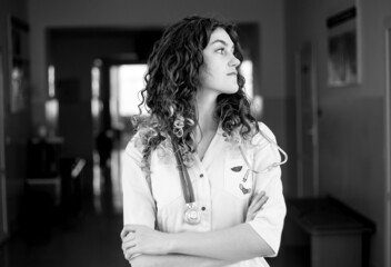 A woman doctor in a protective mask and a white coat stands against the background of a polyclinic black and white photo