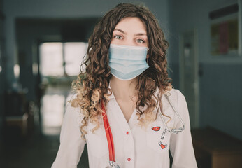 A woman doctor in a protective mask and a white coat stands against the backdrop of a hospital