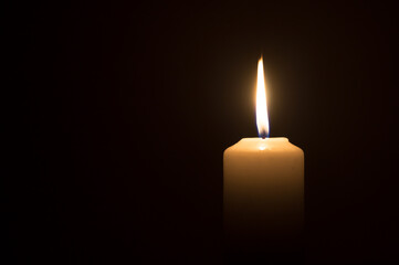 A single burning candle flame or light glowing on a white candle on black or dark background on table in church for Christmas, funeral or memorial service