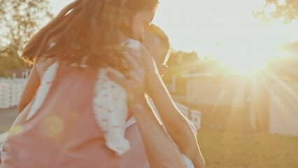 Medium backlighted of happy young Caucasian dad circling beloved daughter in arms outside in sunlight on warm summer day