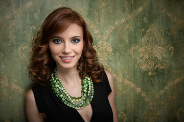 Beautiful young woman with evening make-up and curly hair, with green beads against the background of green wallpaper