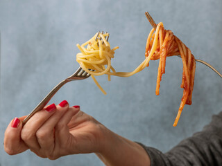 A woman's hands hold two forks with spaghetti in white on one side and spaghetti with tomato sauce...
