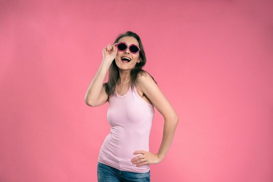 Sexy happy girl in sun glasses kiss shaped wearing tank top and denim jeans posing in studio isolated on pink background