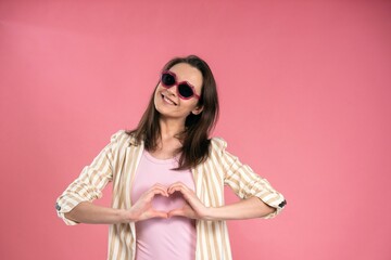  Happy business woman in sun glasses kiss shaped showing love sign with hands shaped in heart, wearing stripe jacket posing in studio isolated on pink background