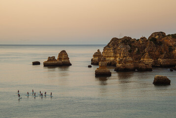SUP touring. Group of tourists on sap boards slowly swim on smooth water to explore local unusual rocks from water. Lagos, Portugal