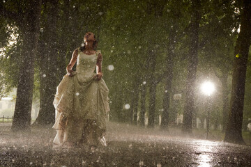 happy bride in a white wedding dress in the rain on an alley under big green trees walking peaying...