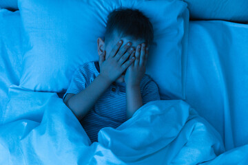 Small boy lies in bed in dark night and covers his face with his hands in fear, afraid of...
