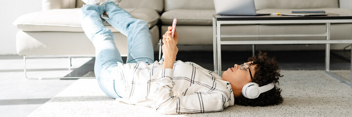 Young curly woman in headphones using cellphone while lying on floor