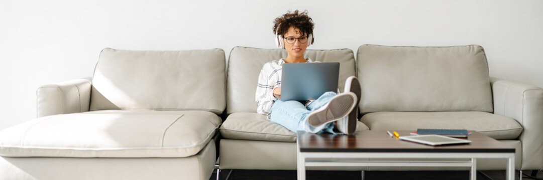 Young Curly Woman In Headphones Using Laptop While Sitting On Couch