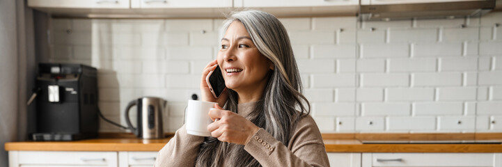 Mature smiling woman talking on cellphone while having breakfast