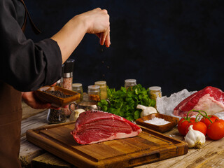 Fototapeta na wymiar Cooking meat dishes by chef's hands on a wooden cutting board on a dark background. Vegetables, herbs, seasonings, spices Lots of objects. Recipes for meat dishes, cookbook, advertising.