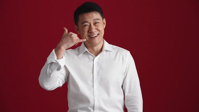 Positive Asian man in white shirt showing call me back gesture to the camera in the red studio