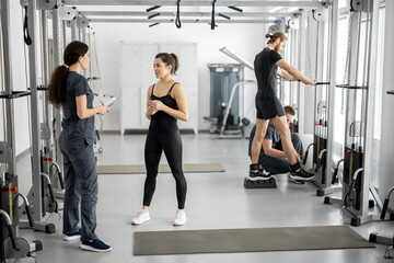 Fototapeta premium People at rehabilitation center, woman talking with female rehabilitologist, men training on background. Concept of physical therapy for health and recovery. Consultation before training