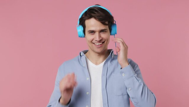 Vivid cheerful calm young brunet man 20s years old wears blue shirt listen music in headphones dance have fun show thumb up like gesture enjoy relax isolated on plain pink background studio portrait